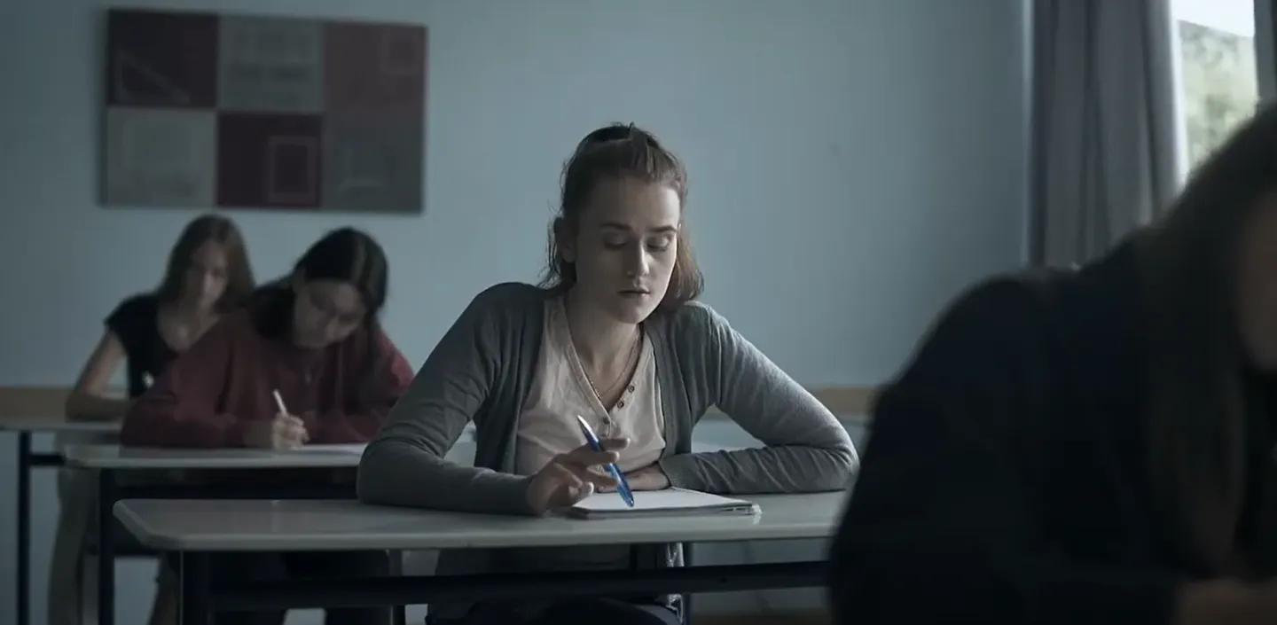 Eine junge Frau sitzt an einem Schreibtisch in einem Klassenraum und hält einen blauen Kugelschreiber über ein leeres Blatt Papier.