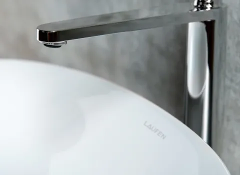 Ein modernes, weißes Waschbecken der Marke "Laufen" steht unter einem eleganten, verchromten Wasserhahn. Der Hahn hat ein minimalistisches Design mit einem langen, schmalen Auslauf, der sich über das Waschbecken erstreckt.