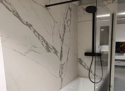 Badewanne mit Glasabtrennung und schwarzem Duschsystem vor marmorierter Wand in einer zeitgemäßen Badezimmerausstellung.